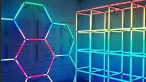A geometric light installation features glowing neon hexagons and cubes in vibrant rainbow colors, illuminating a dark room with colorful reflections on the walls.