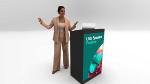 A woman in a beige suit stands at a podium labeled LED Speaker Podium, gesturing with one hand while speaking. The podium features a colorful abstract design on the front.
