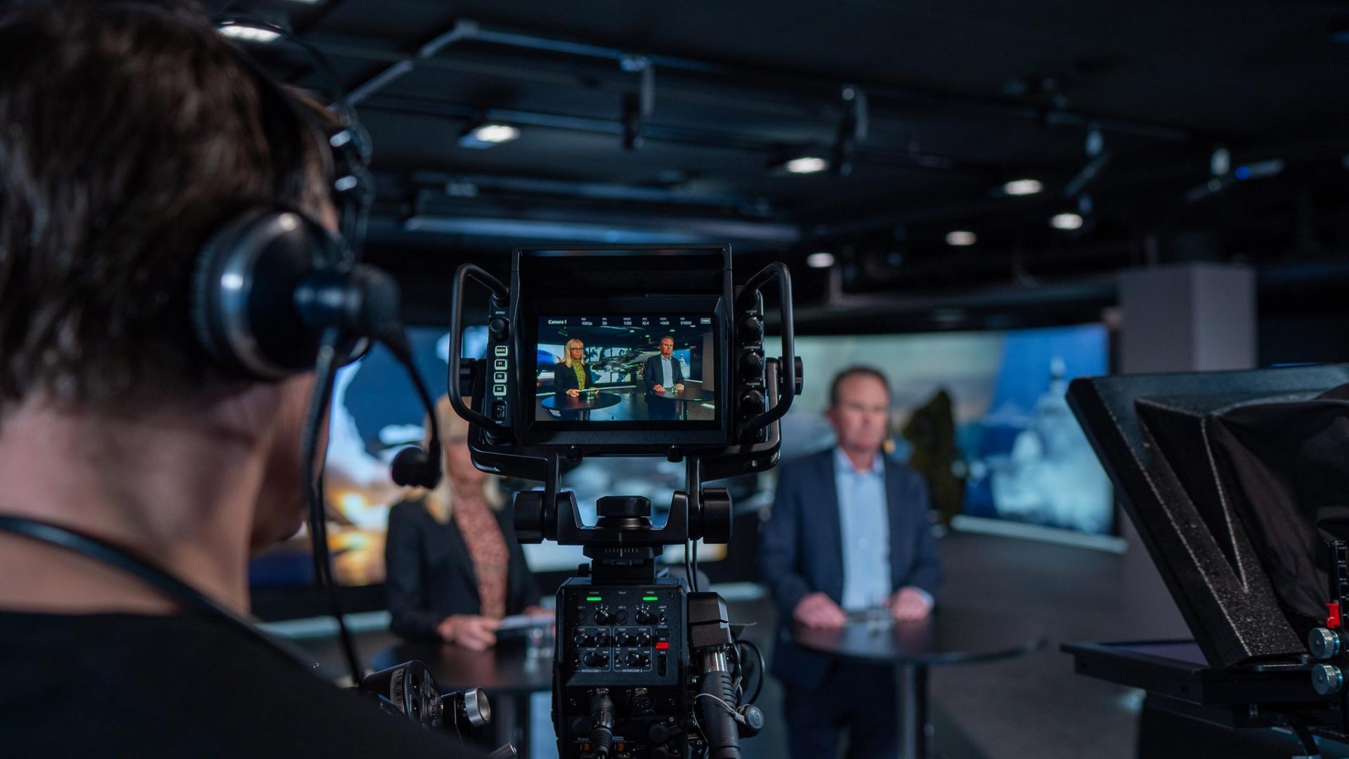 A camera operator films two people in a studio, one man and one woman, who are standing at a news desk. The camera screen shows them clearly while the background is blurred. Studio lights and equipment are visible.