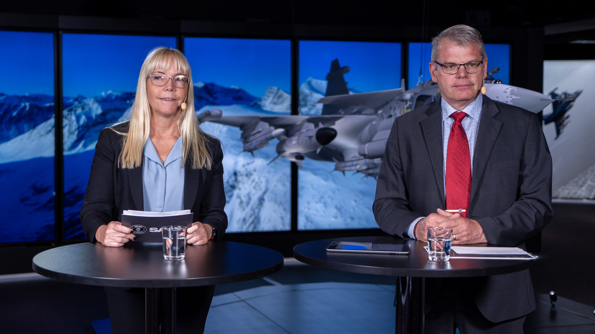A woman and a man in business attire stand at separate round tables with glasses of water, holding notepads. Behind them is a large screen showing a snowy mountain and a fighter jet.