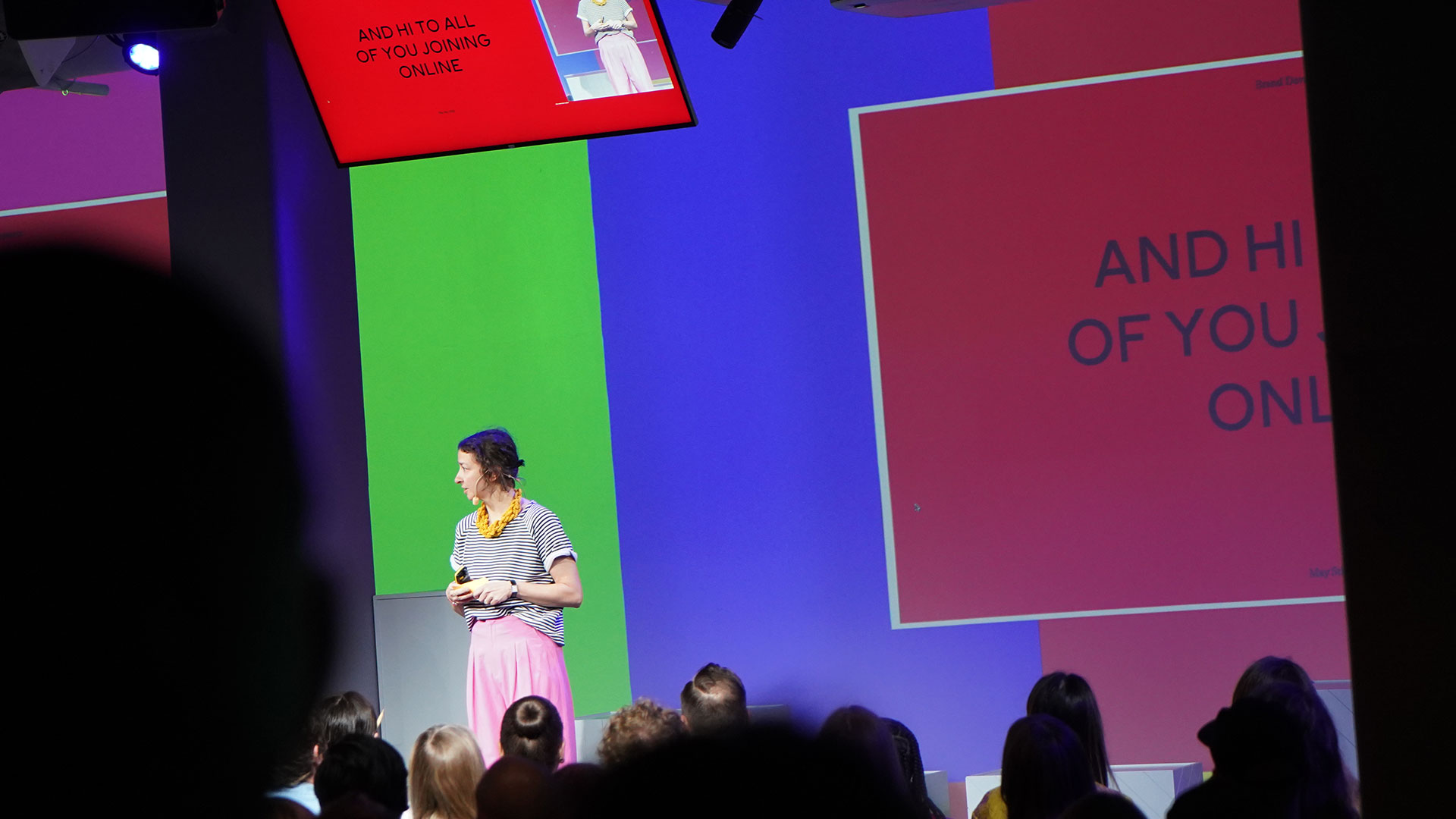 A speaker in a striped shirt and pink skirt stands on stage addressing an audience, with colorful screens behind her displaying the words AND HI TO ALL OF YOU JOINING ONLINE.