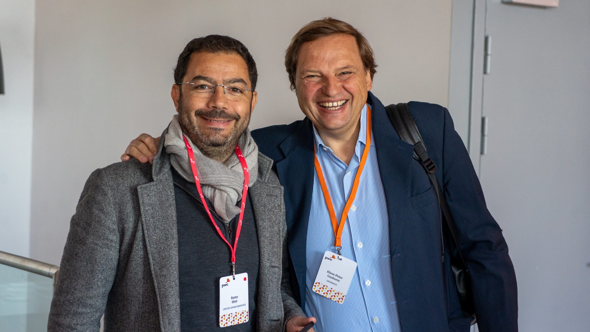 Two men smiling and posing together indoors. Both wear conference badges; one is in a gray coat and scarf, the other in a navy blazer with an orange lanyard and backpack. They appear friendly and relaxed.