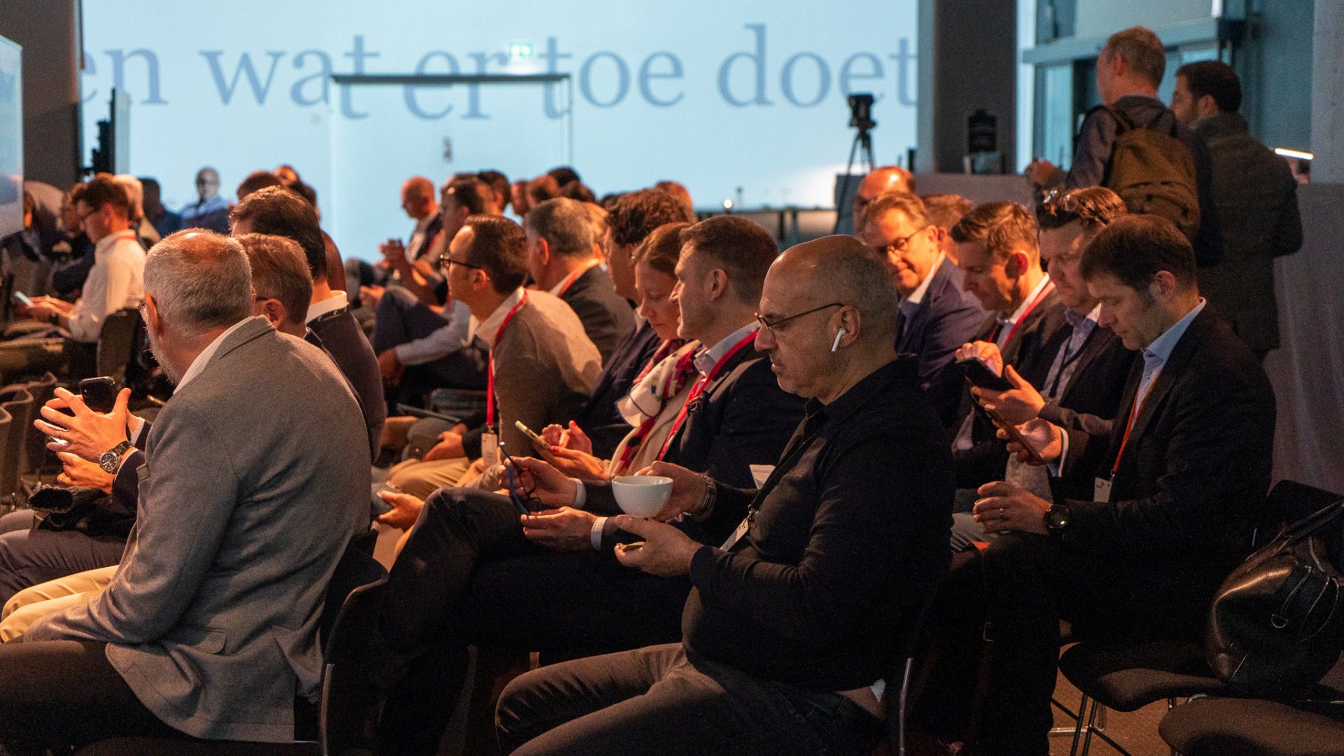 A group of people in business attire sit in rows at a conference, some using their phones or drinking coffee. The background features large windows and projected text on the wall.