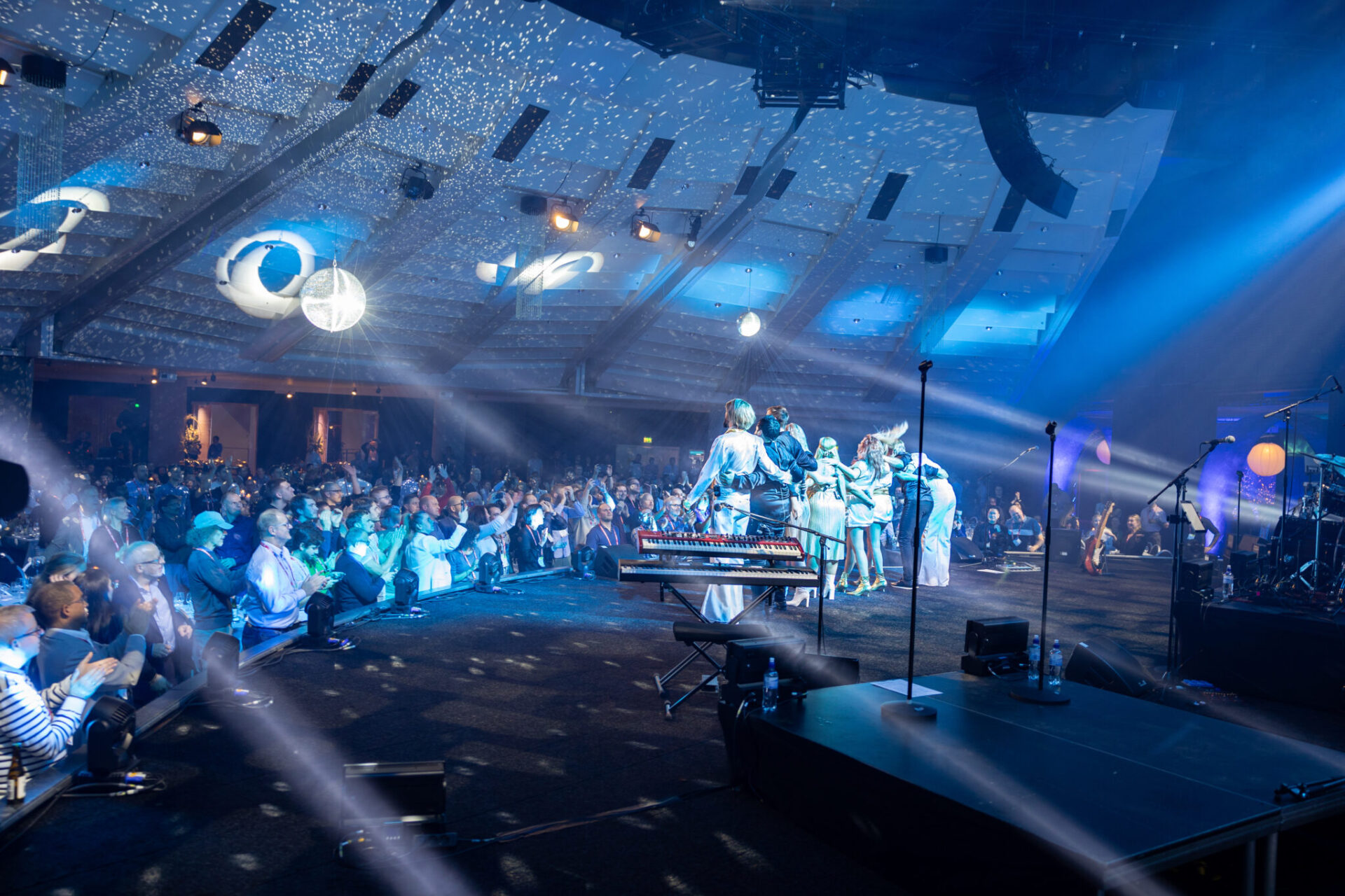 A musical group performs on a brightly lit stage before a large seated audience in a spacious venue, with blue lights and star-like patterns projected onto the ceiling and walls.