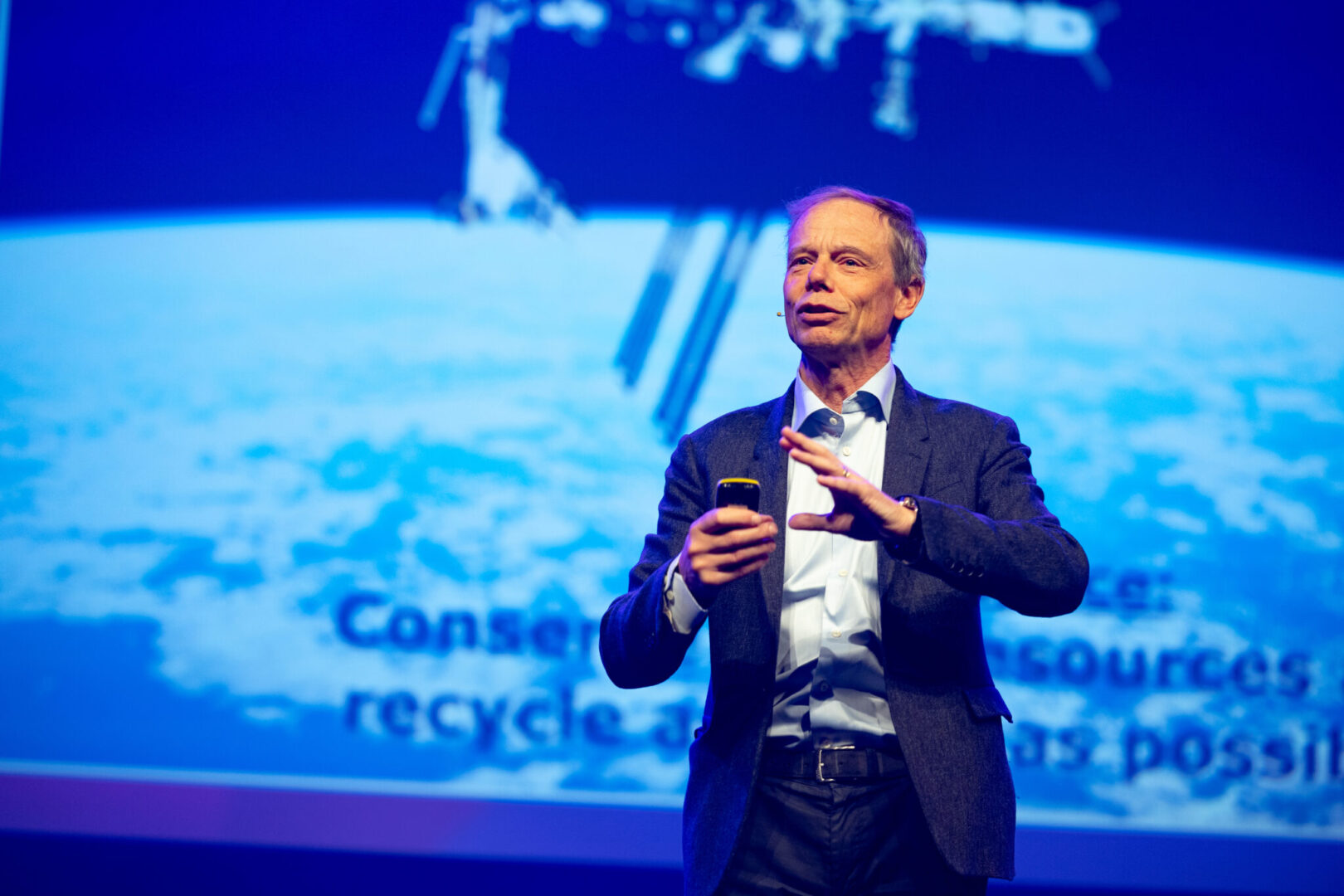 A man in a suit speaks on stage holding a device, with a large screen behind him displaying an image of Earth from space and partially visible text.