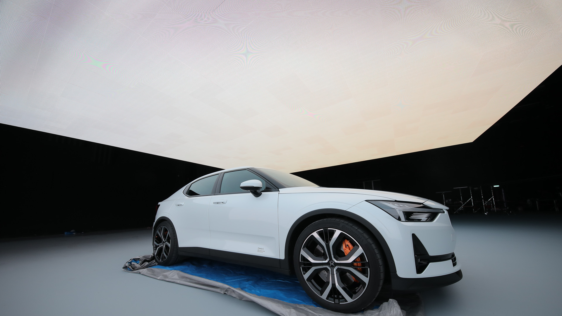 A white modern electric car parked indoors under a large, illuminated ceiling panel, with a plastic sheet partially underneath the vehicle. The background is dark and the scene appears to be part of a photoshoot or display.