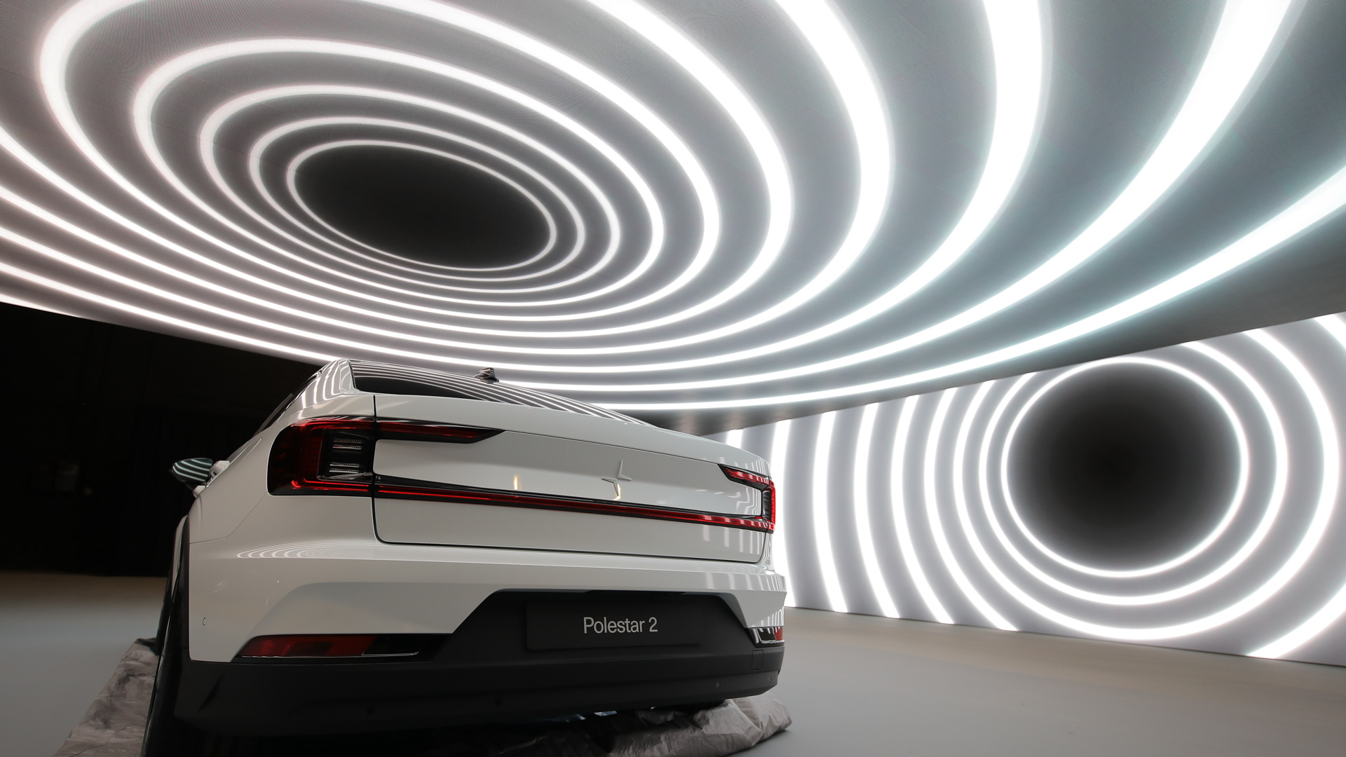A white Polestar 2 car is displayed indoors, with bright, concentric circular light patterns on the ceiling and walls creating a futuristic atmosphere around the vehicle.