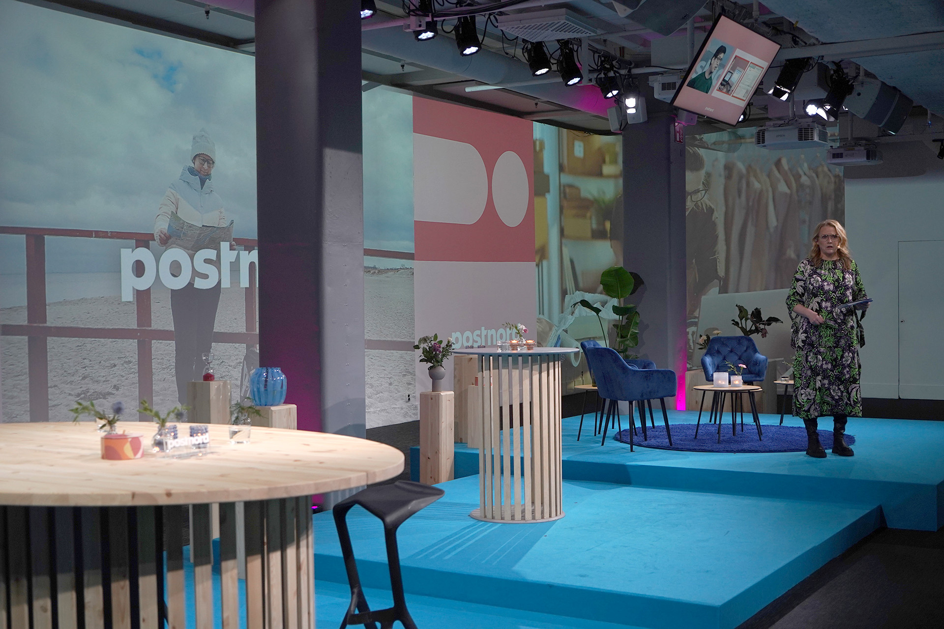 A woman stands on a blue stage in a modern studio with round wooden tables, chairs, and plants. Large screens behind her display the word “postnord” and various images. The lighting is purple and blue.