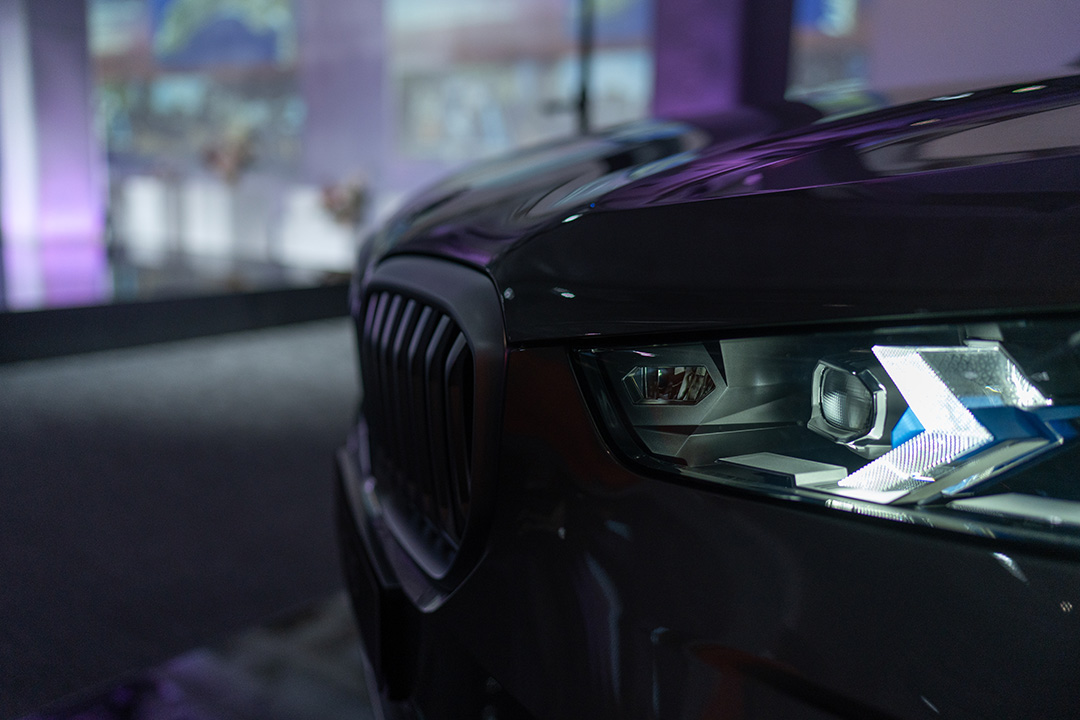 Close-up of the front left side of a dark-colored car, focusing on the headlight and part of the grille, with a blurred background featuring soft purple lighting.