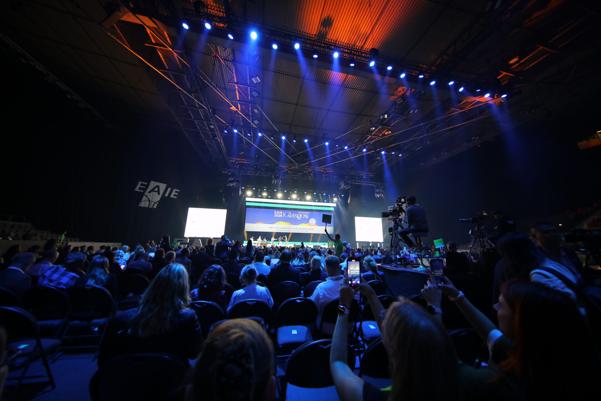A large audience watches a brightly lit stage with blue lights at an indoor event. People are seated, some taking photos with their phones. The backdrop reads EAIE and features a central screen with presentations.