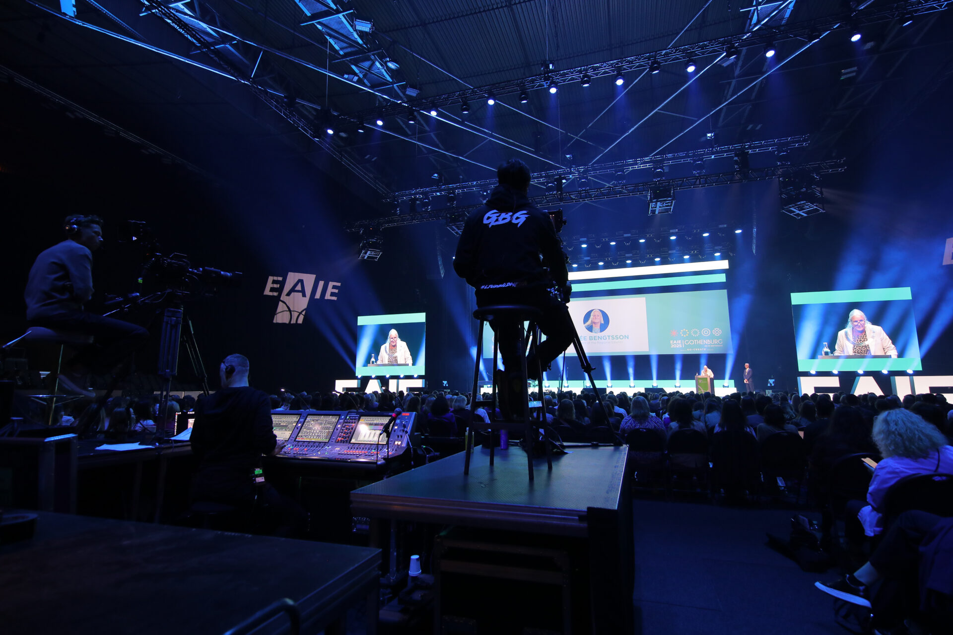 A large conference or event with an audience seated in front of a stage, where a presenter speaks at a podium. Camerapersons and technical staff are visible in the foreground, capturing and managing the event.