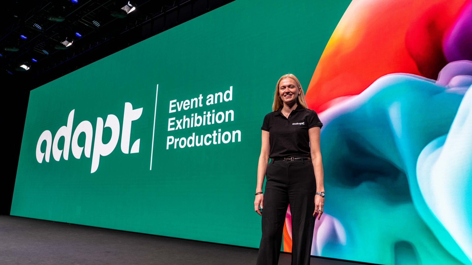 A woman in black stands smiling in front of a large screen displaying the words adapt | Event and Exhibition Production with colorful abstract graphics on the right side.