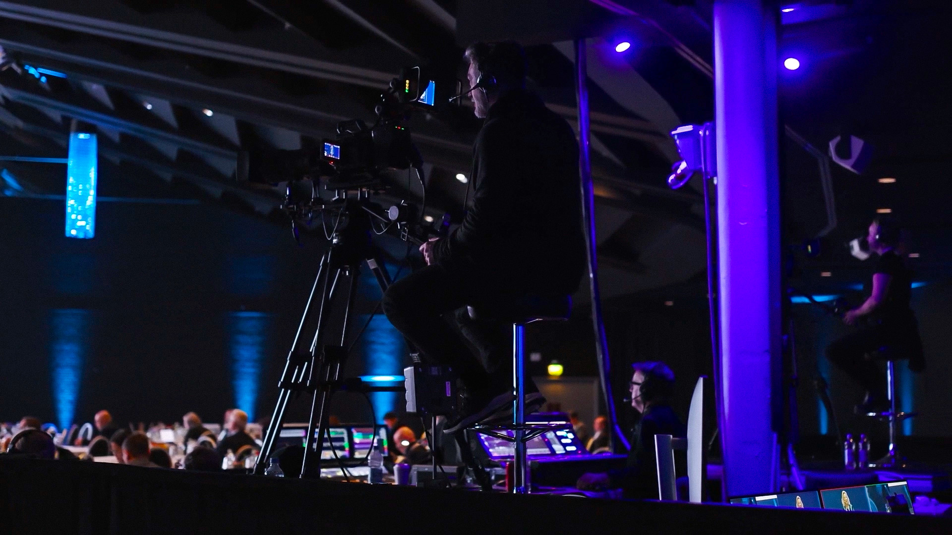A camera operator sits on a tall stool filming an event in a dimly lit auditorium with blue lighting, while attendees are seated in the background.