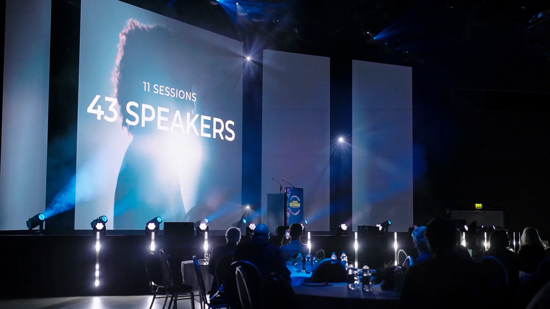 A dimly lit conference hall with people seated at round tables, facing a stage with a large screen displaying “11 sessions, 43 speakers.” Spotlights illuminate the stage, and a podium stands in front of the screen.
