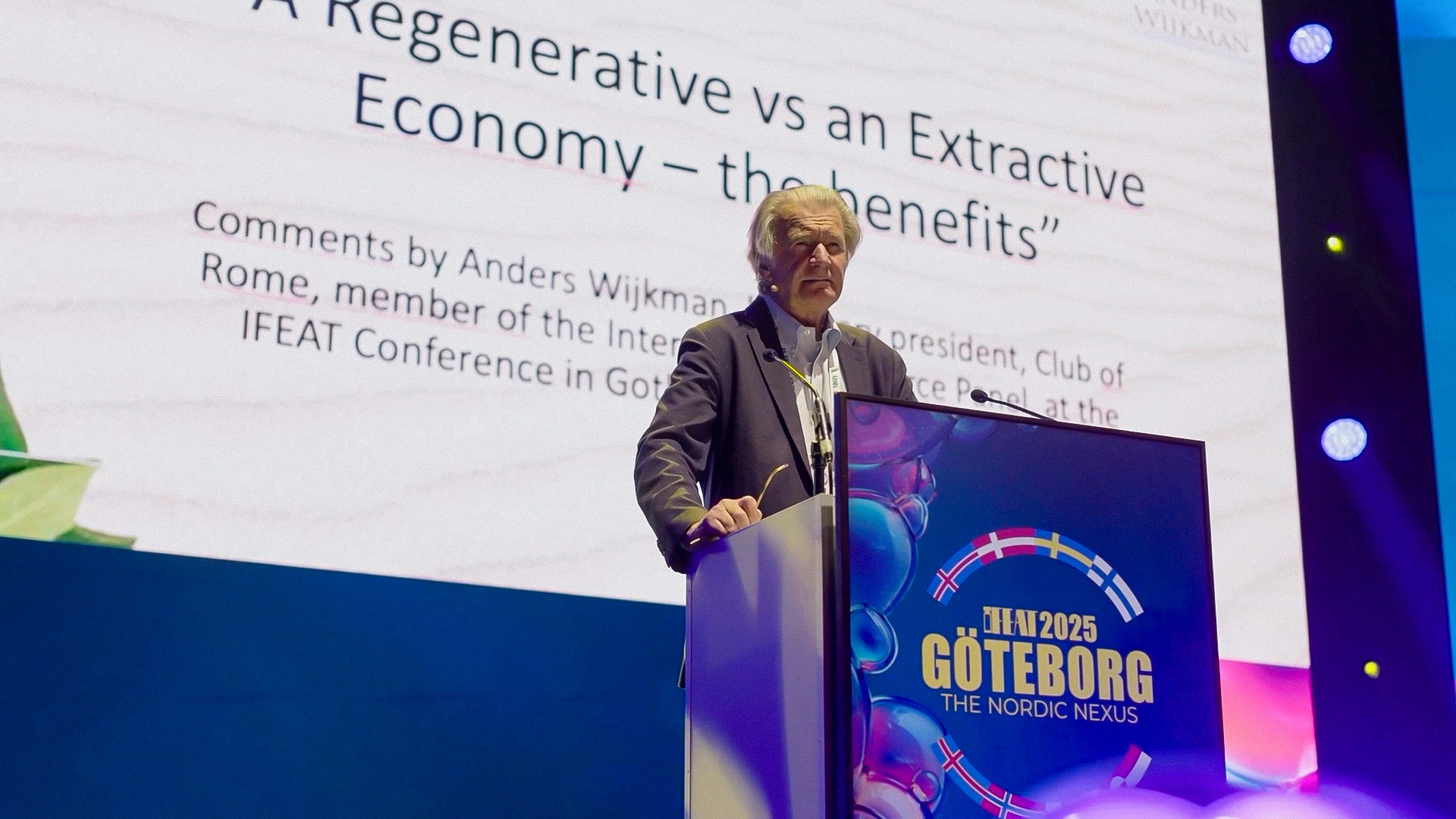 An older man stands at a podium labeled I FEAT 2025 GÖTEBORG THE NORDIC NEXUS, delivering a presentation titled A Regenerative vs an Extractive Economy – the benefits, with a large screen behind him.