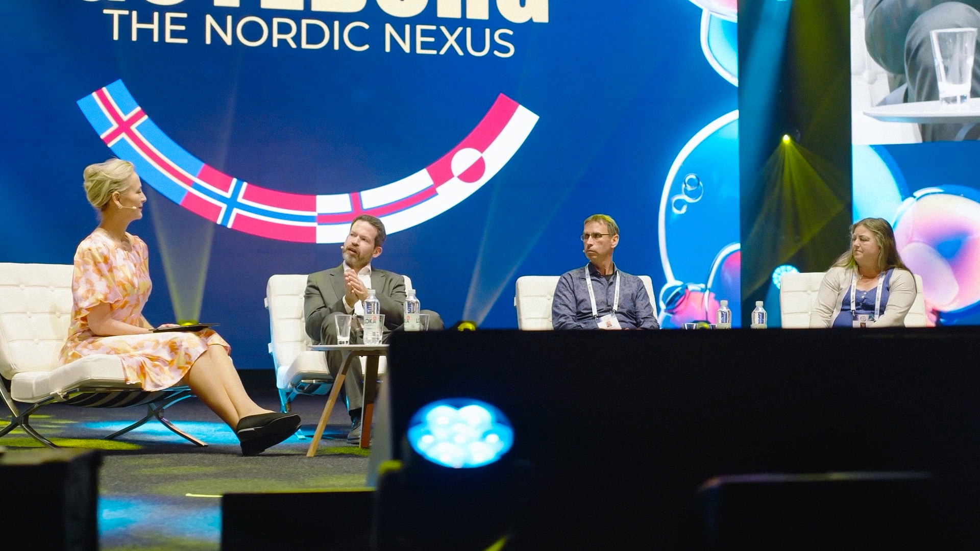 Four people sit on a stage having a panel discussion, with a large screen behind them displaying THE NORDIC NEXUS and Nordic flag designs. The participants are seated in white chairs with microphones.