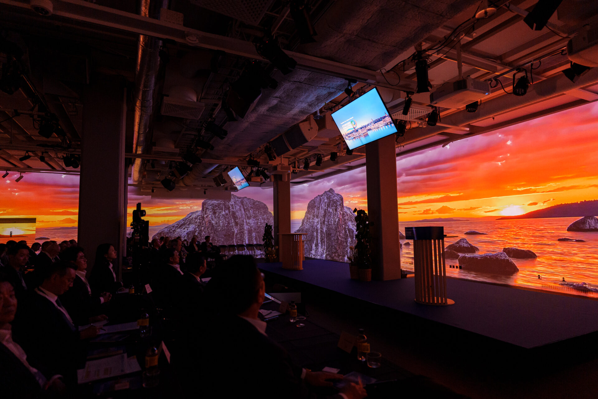 A conference room with people seated in the audience, facing a stage. Large digital screens display a vivid sunset over rocky islands and sea, creating an immersive backdrop. The atmosphere is formal and modern.