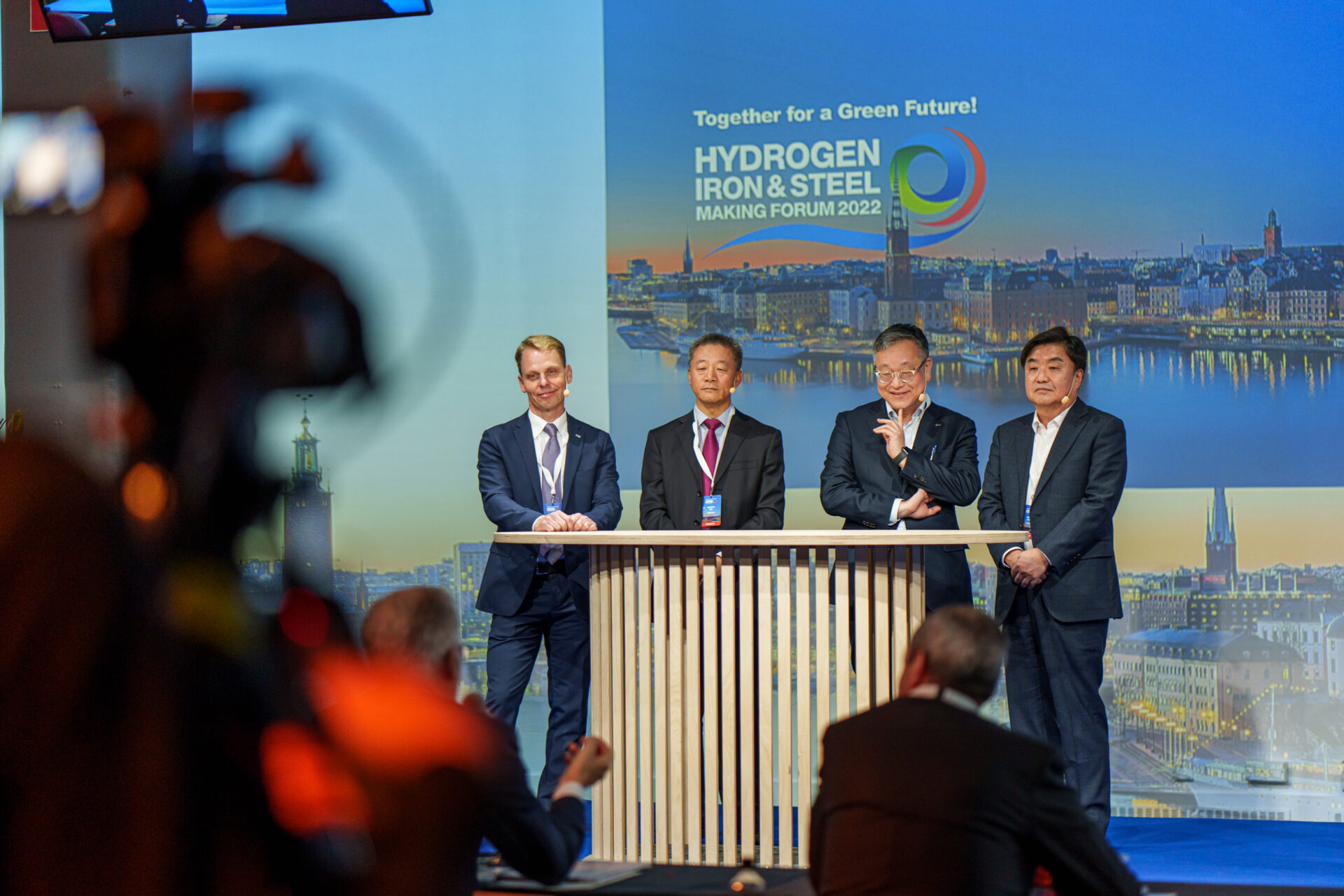 Four men in business attire stand behind a podium on stage at the Hydrogen Iron & Steel Making Forum 2022, with a blue backdrop and cityscape image, as photographers and a camera capture the event.