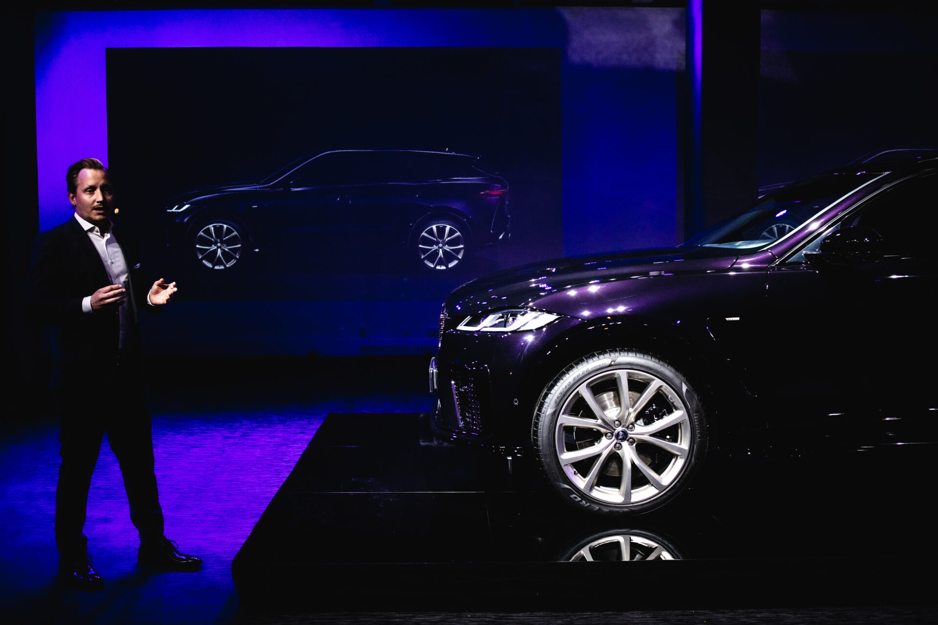 A man in a suit stands on stage next to a shiny black car, with a side profile image of the same car projected on a screen behind him, under blue and purple lighting.