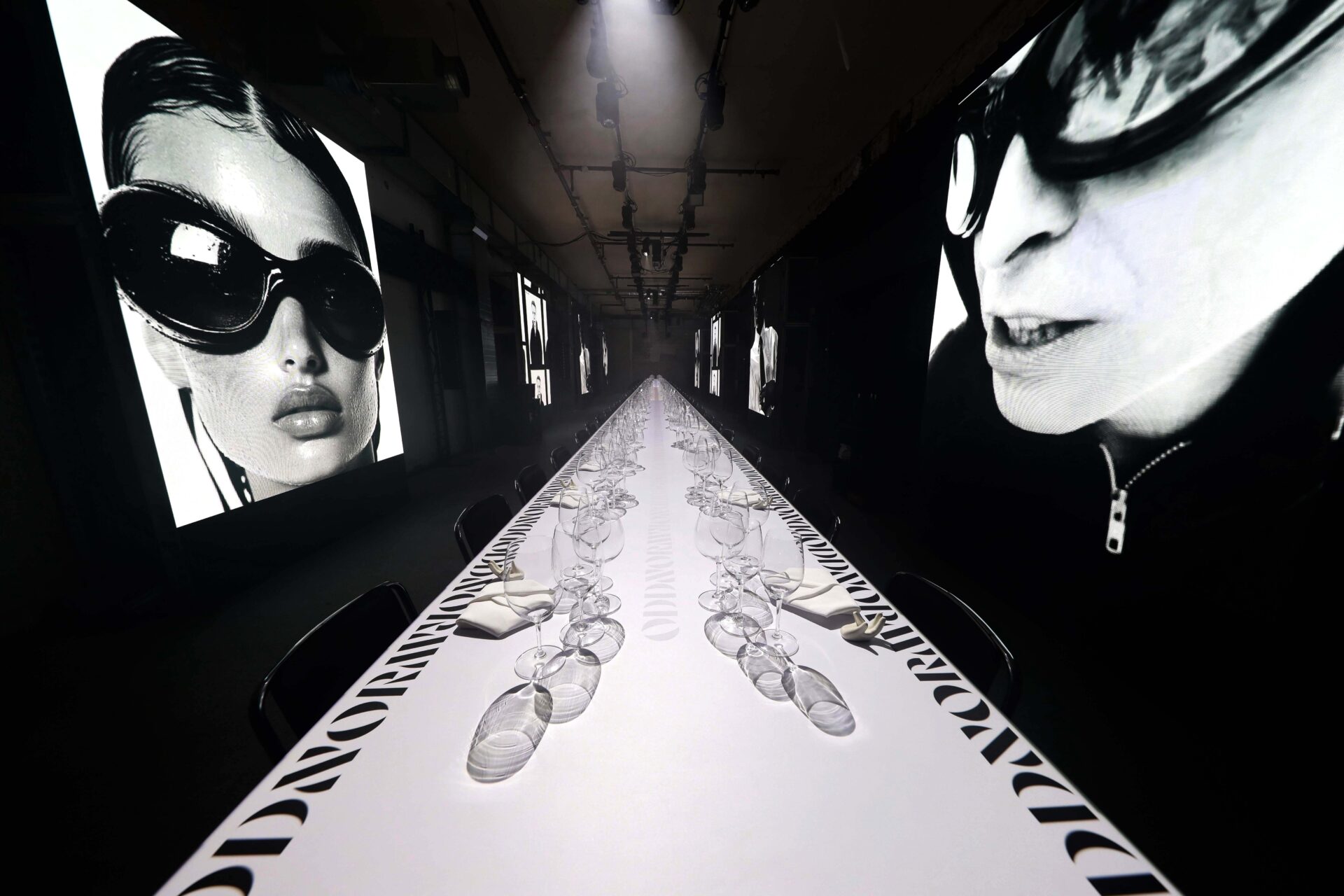 A long, white runway-style table is set with glassware and surrounded by black chairs. Large black-and-white portraits of people in sunglasses are displayed on screens along the dark walls.