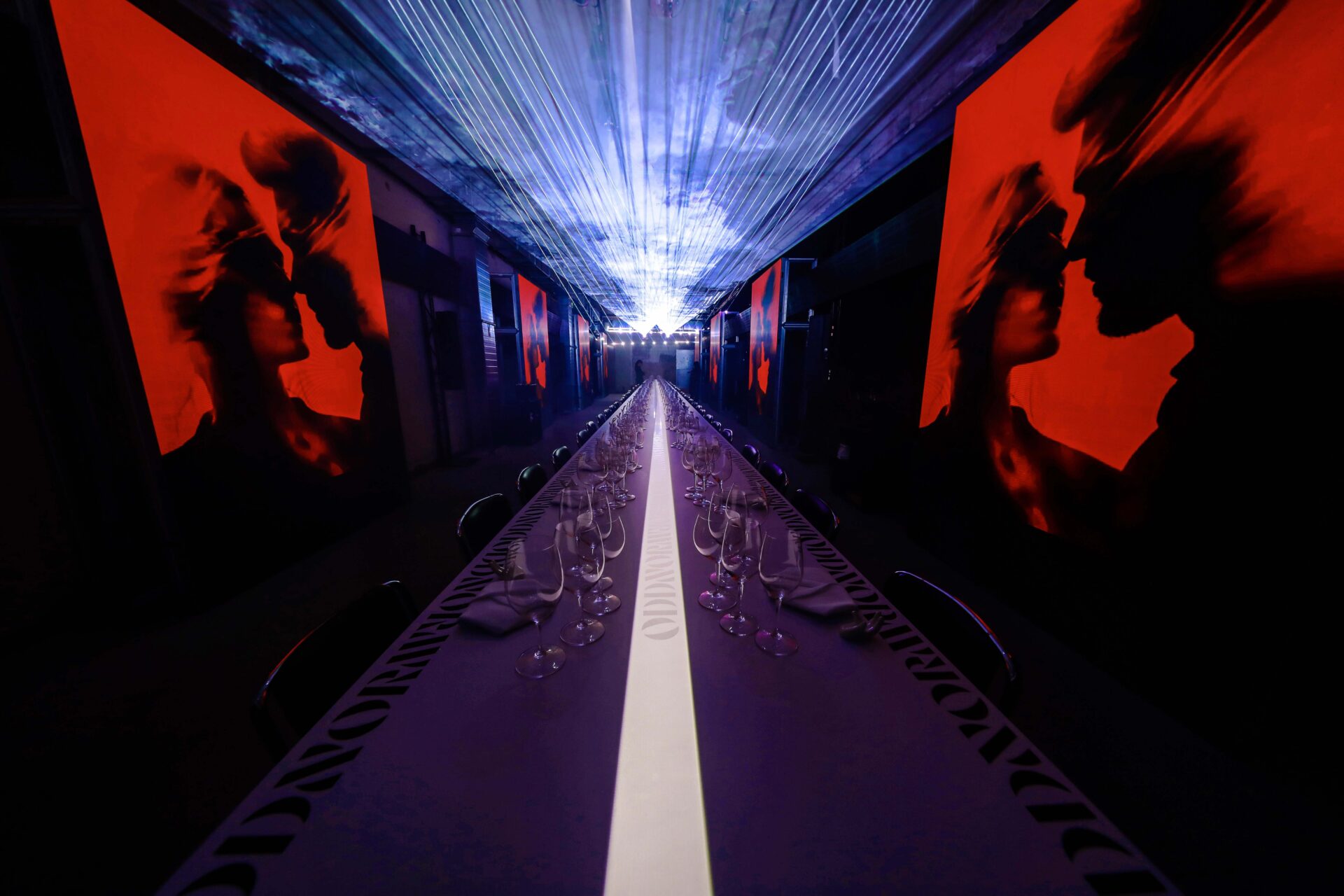 A long, elegant dining table is set with empty wine glasses, beneath dramatic lighting and a ceiling of reflective material. Large red and black silhouette art pieces depicting couples face each other along the walls.