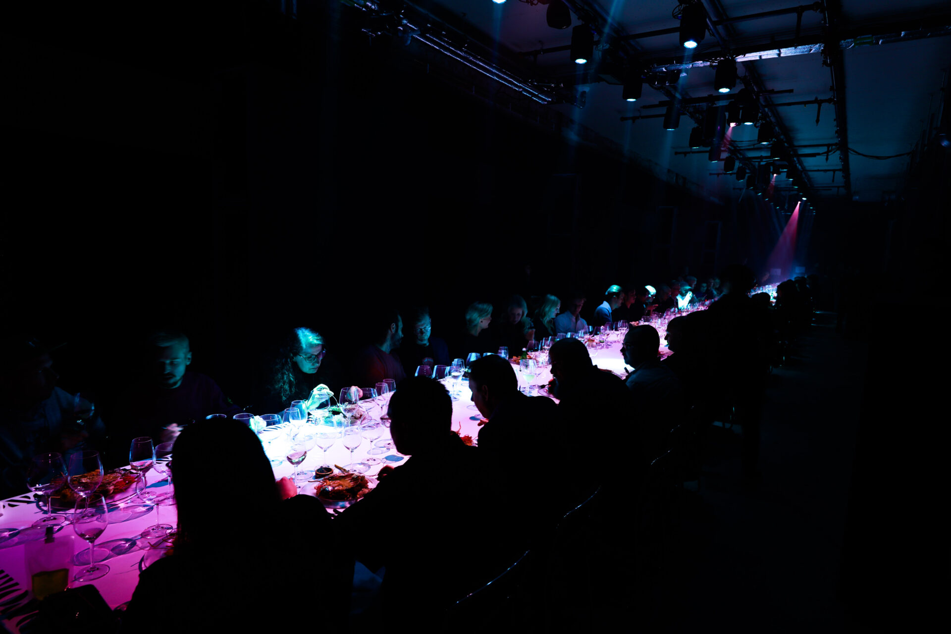 A long dining table with people seated on both sides under dim, colorful lighting, creating a dramatic and atmospheric scene in a dark room. Glasses and plates are visible on the table.