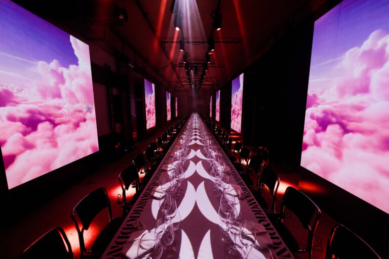 A long dining table set with glassware and surrounded by chairs is illuminated by dramatic red lighting, with large screens displaying pink clouds lining both sides of the room.