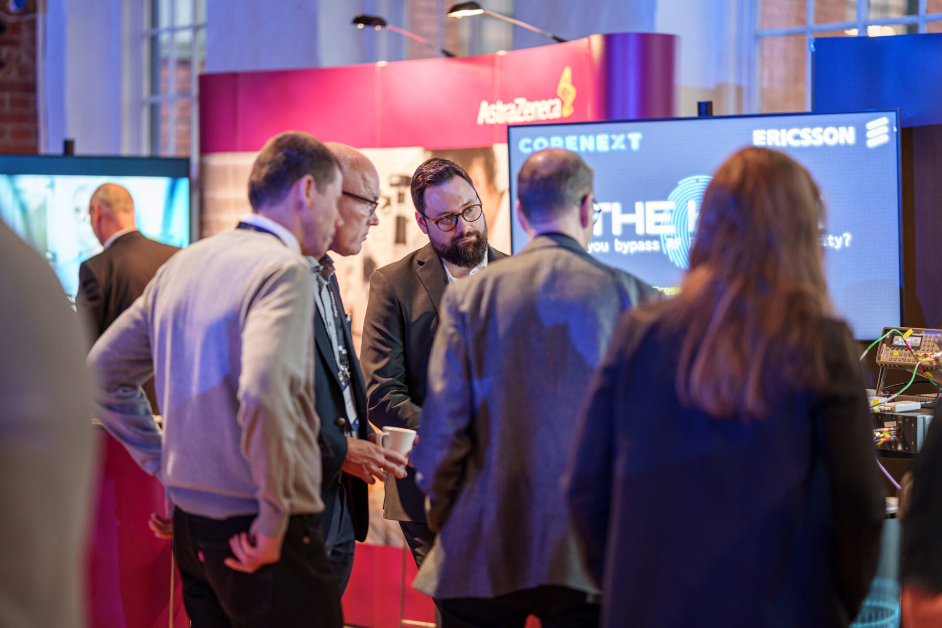 A group of men stand and converse in front of technology company booths, including AstraZeneca and Ericsson, at a professional event or conference. One man appears to be explaining something to the others.