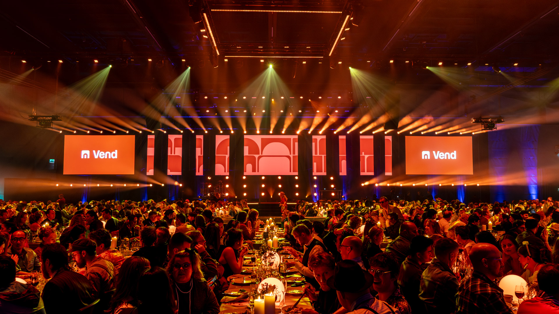 A large crowd sits at decorated tables in a spacious, dimly lit event hall with warm lighting, focused on a brightly lit stage displaying Vend on large screens.