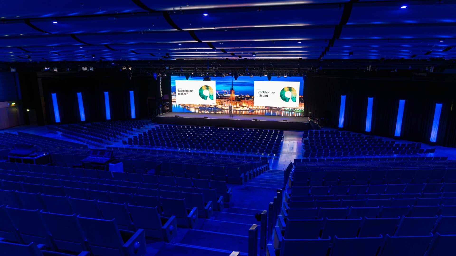 A large, empty auditorium with rows of blue seats facing a stage, which features two large screens displaying the Stockholm+50 logo and an illuminated cityscape in the background. The space is lit with blue lighting.