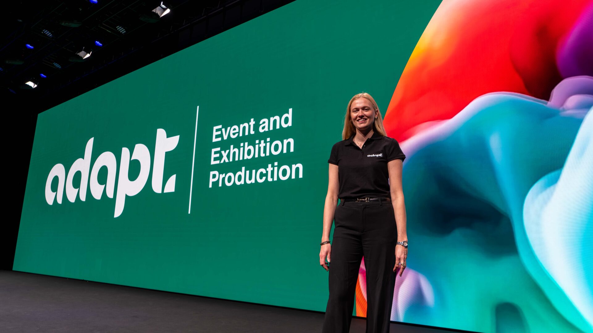 A woman in black business attire stands smiling on stage in front of a large colorful screen displaying the words “adapt Event and Exhibition Production.”.