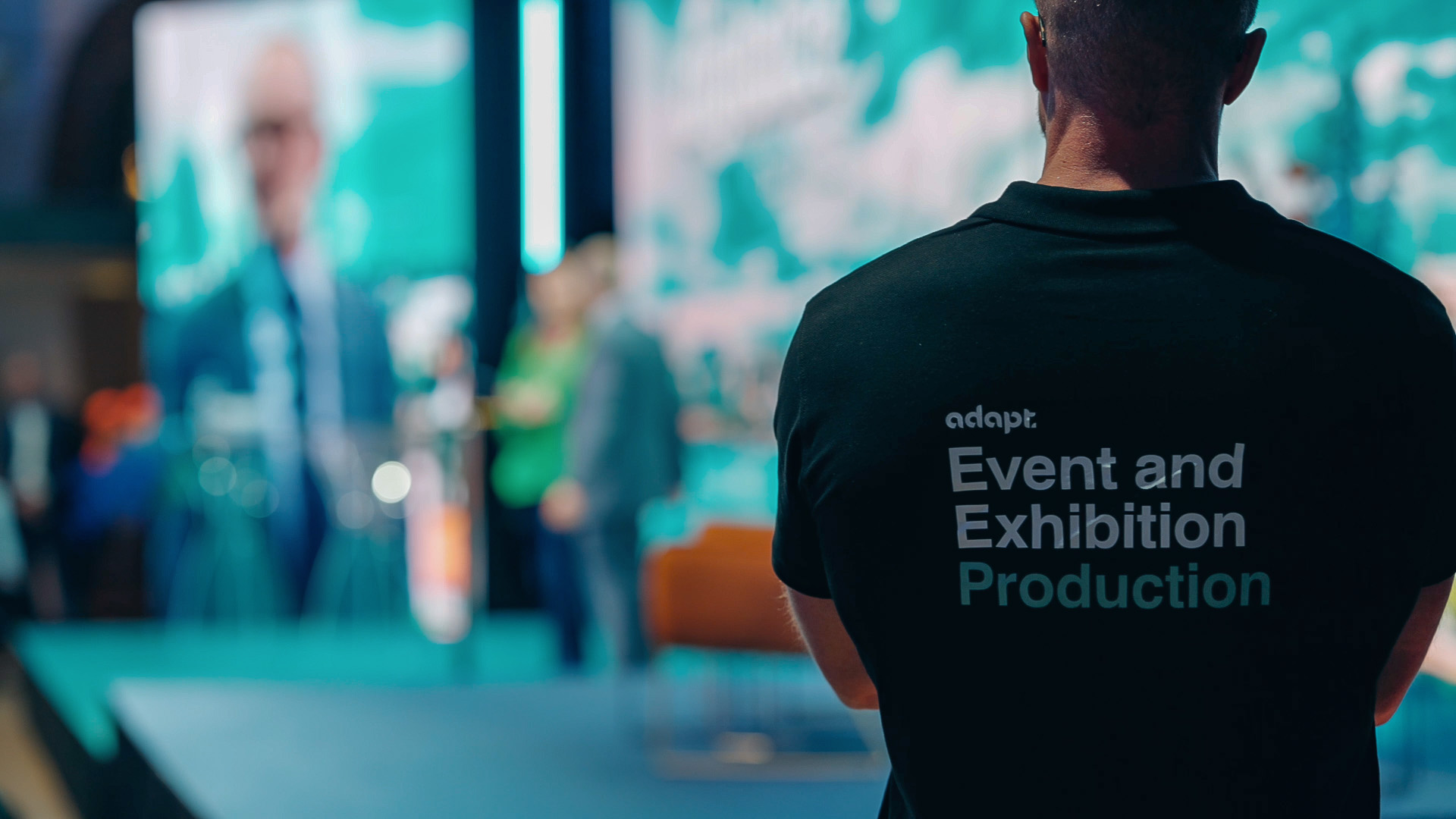 A person wearing a black shirt with Event and Exhibition Production written on the back stands in front of a blurred stage with people and blue lighting in the background.