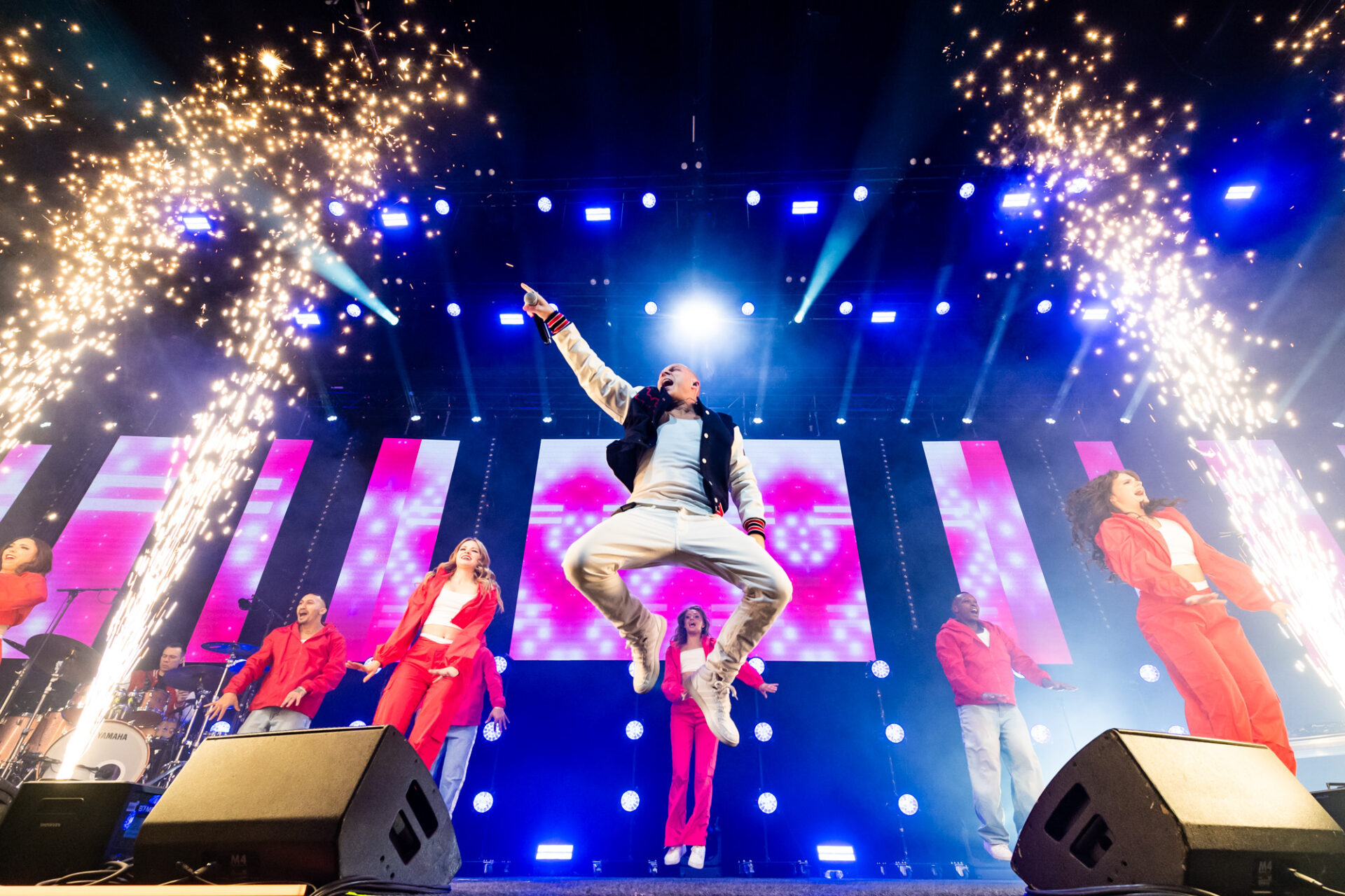 A performer jumps energetically on stage surrounded by backup dancers in red outfits, with bright stage lights, colorful screens, and fireworks effects enhancing the lively concert atmosphere.