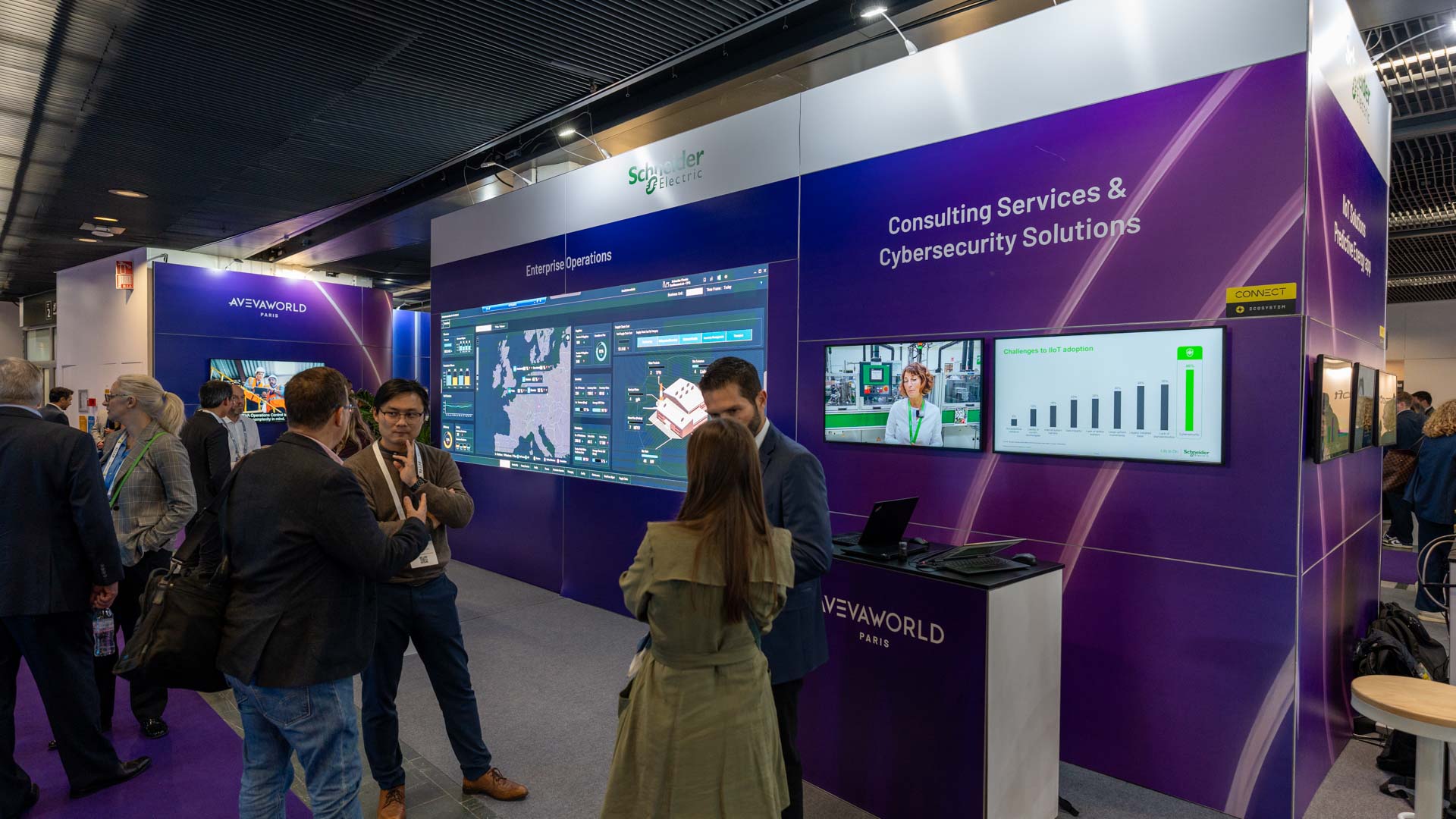 People converse and view digital displays at a tech expo booth for Schneider Electric, showcasing consulting and cybersecurity services. Screens show software dashboards and analytics. The booth features modern design and informational graphics.