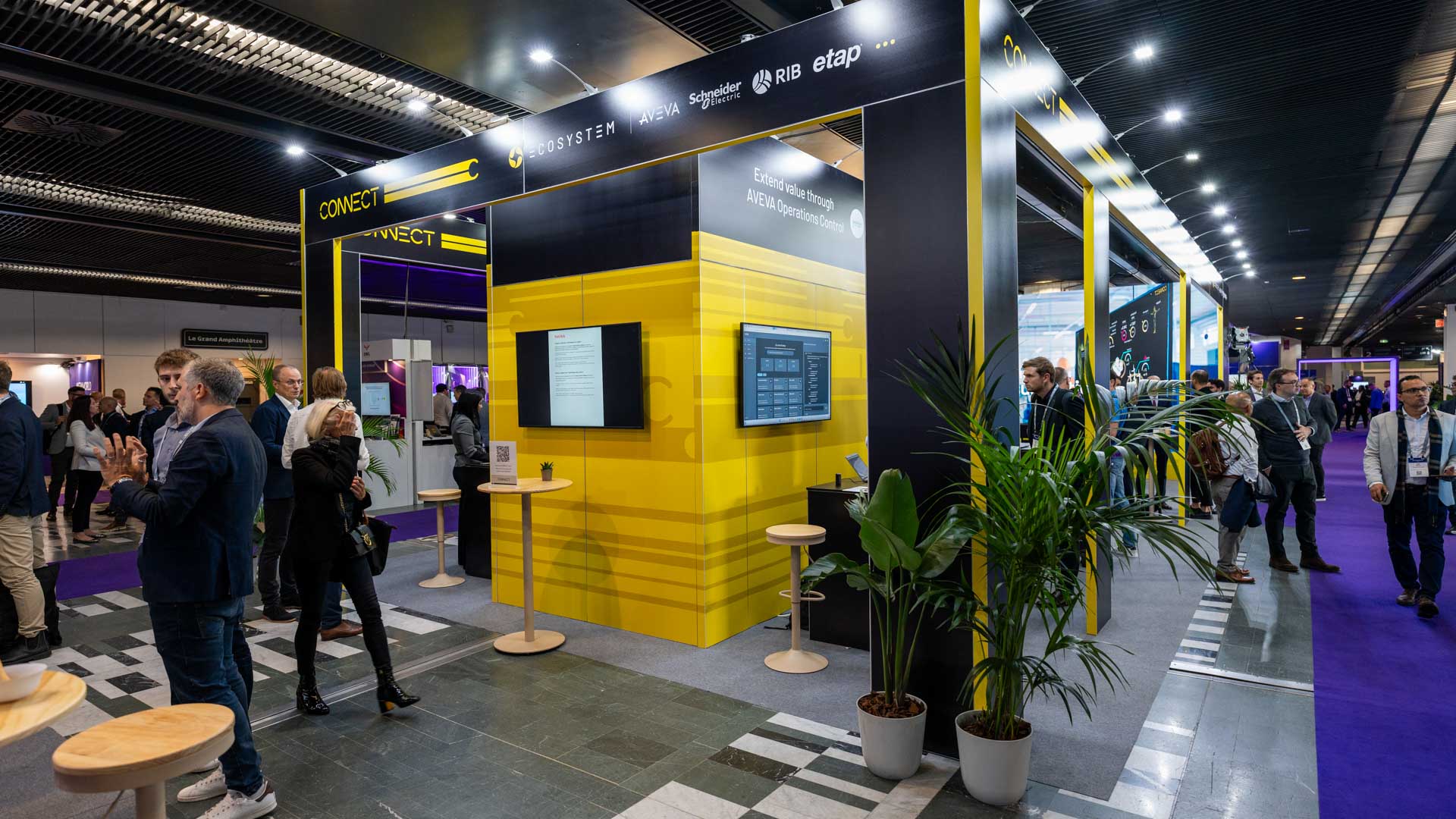 A busy exhibition hall with people interacting near a modern, yellow and black trade show booth featuring digital screens and branded displays, surrounded by plants and high tables.