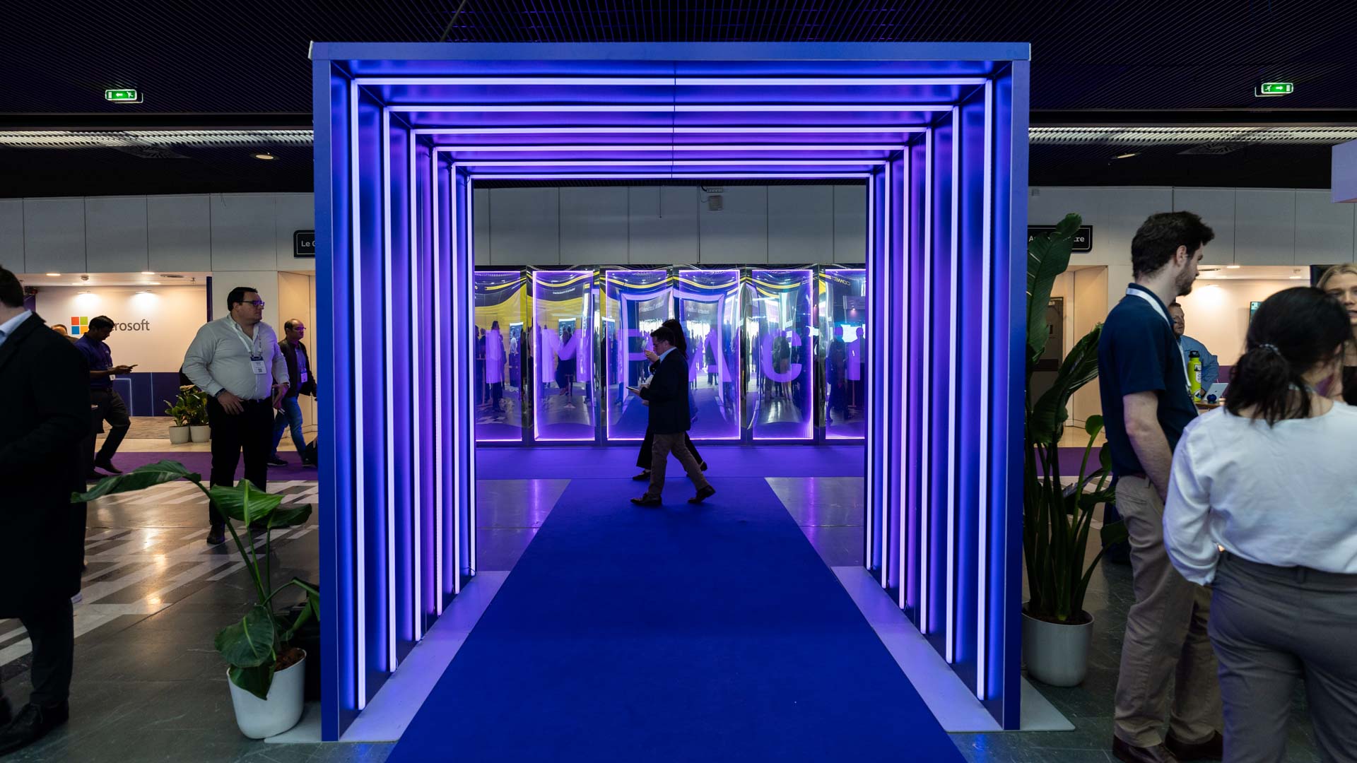 A person walks through a tunnel of vertical blue and purple LED lights at an indoor event, with people standing on either side and booths visible in the background.