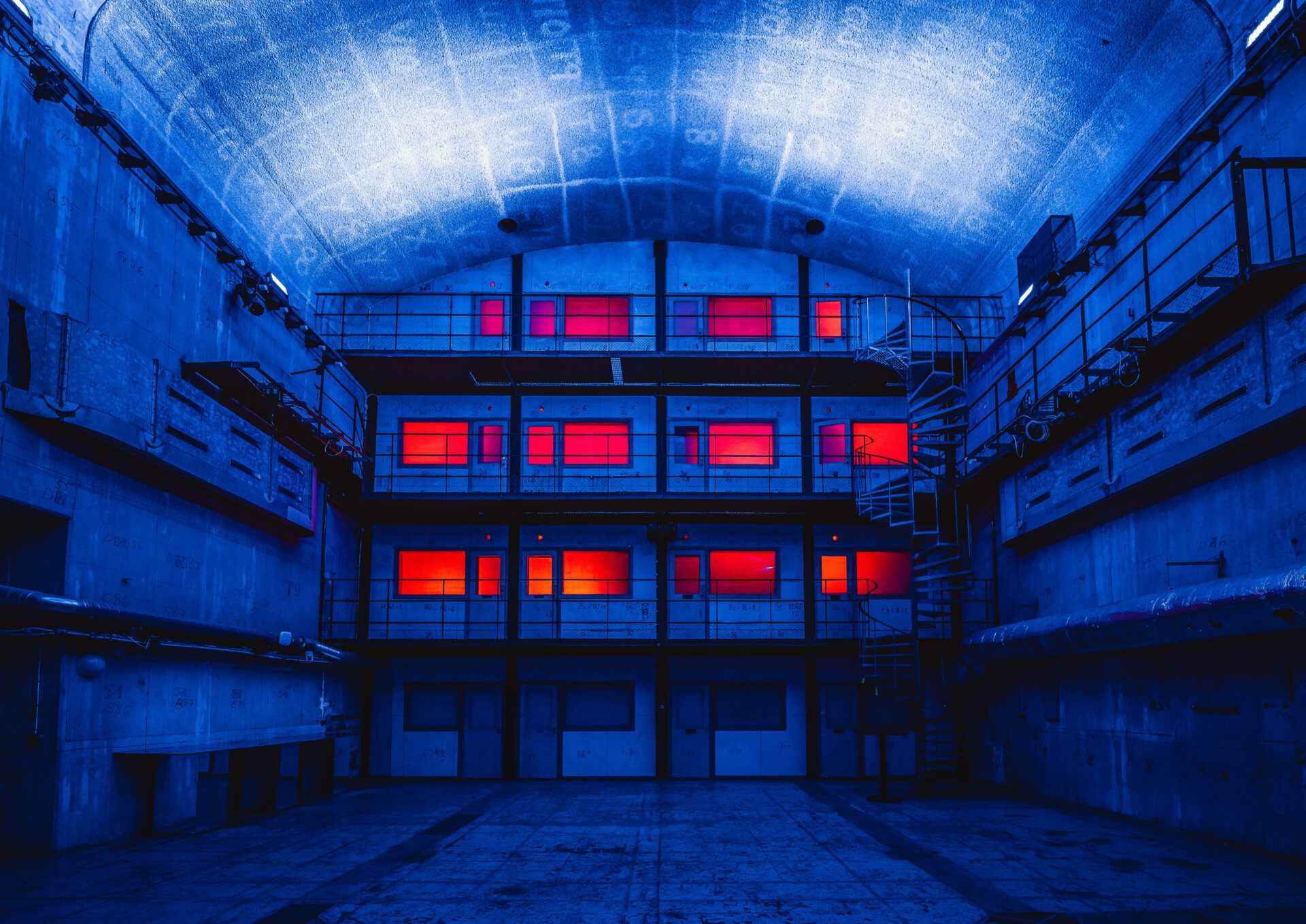 A futuristic, industrial interior with blue-lit walls and ceiling, featuring three floors of rooms with vivid red-lit windows and a spiral staircase on the right side. The setting is empty and atmospheric.