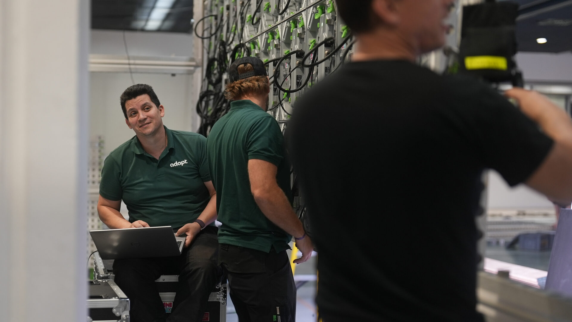 Three men in green shirts work in a tech environment; one sits on a bench with a laptop, smiling at the camera, while the others are standing and working with cables on a large panel.