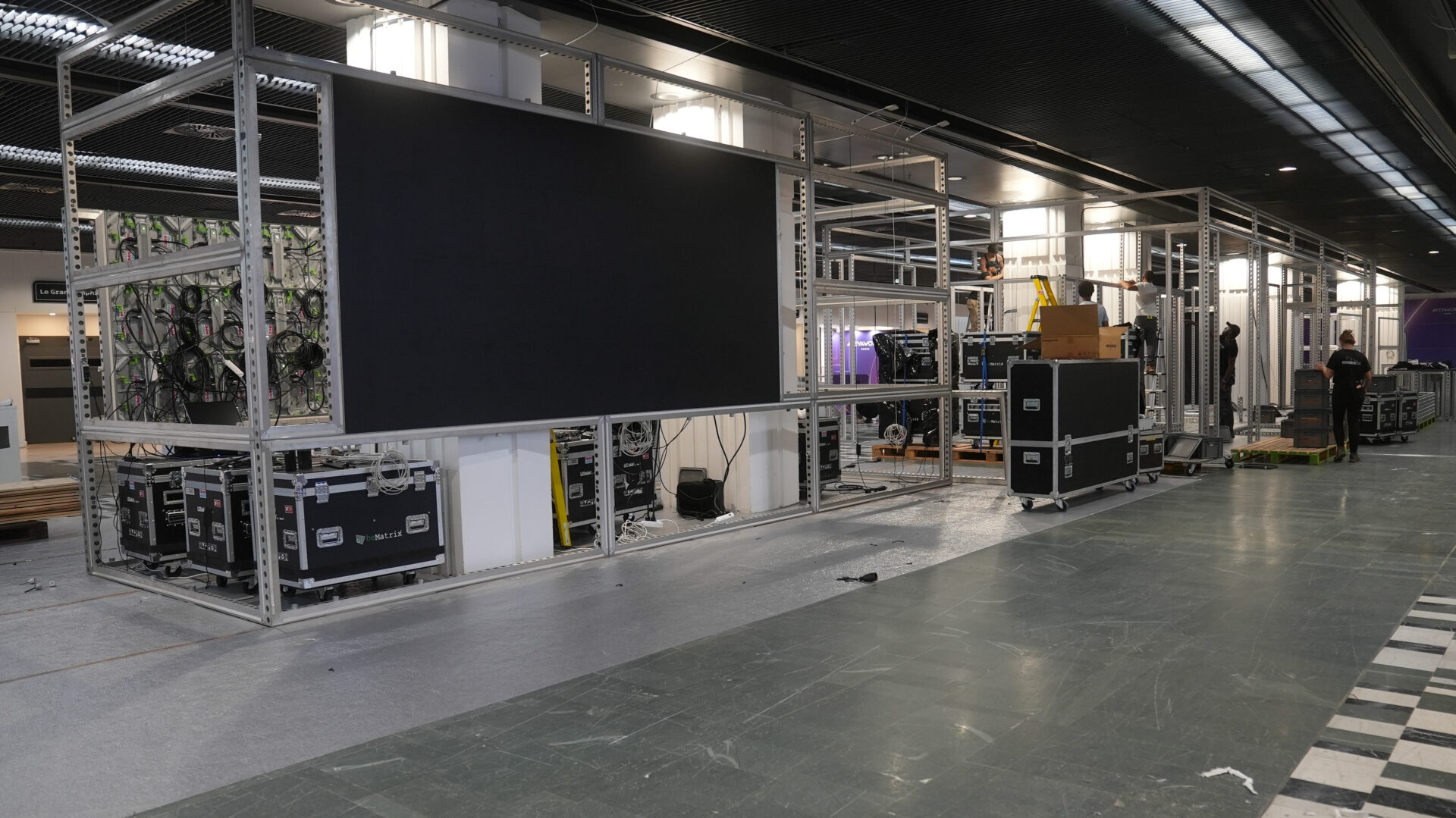 A large indoor space with metal frames, electronic equipment cases, and workers assembling structures, possibly setting up for an event or exhibition. A large screen is mounted on one frame. The floor is partly covered.