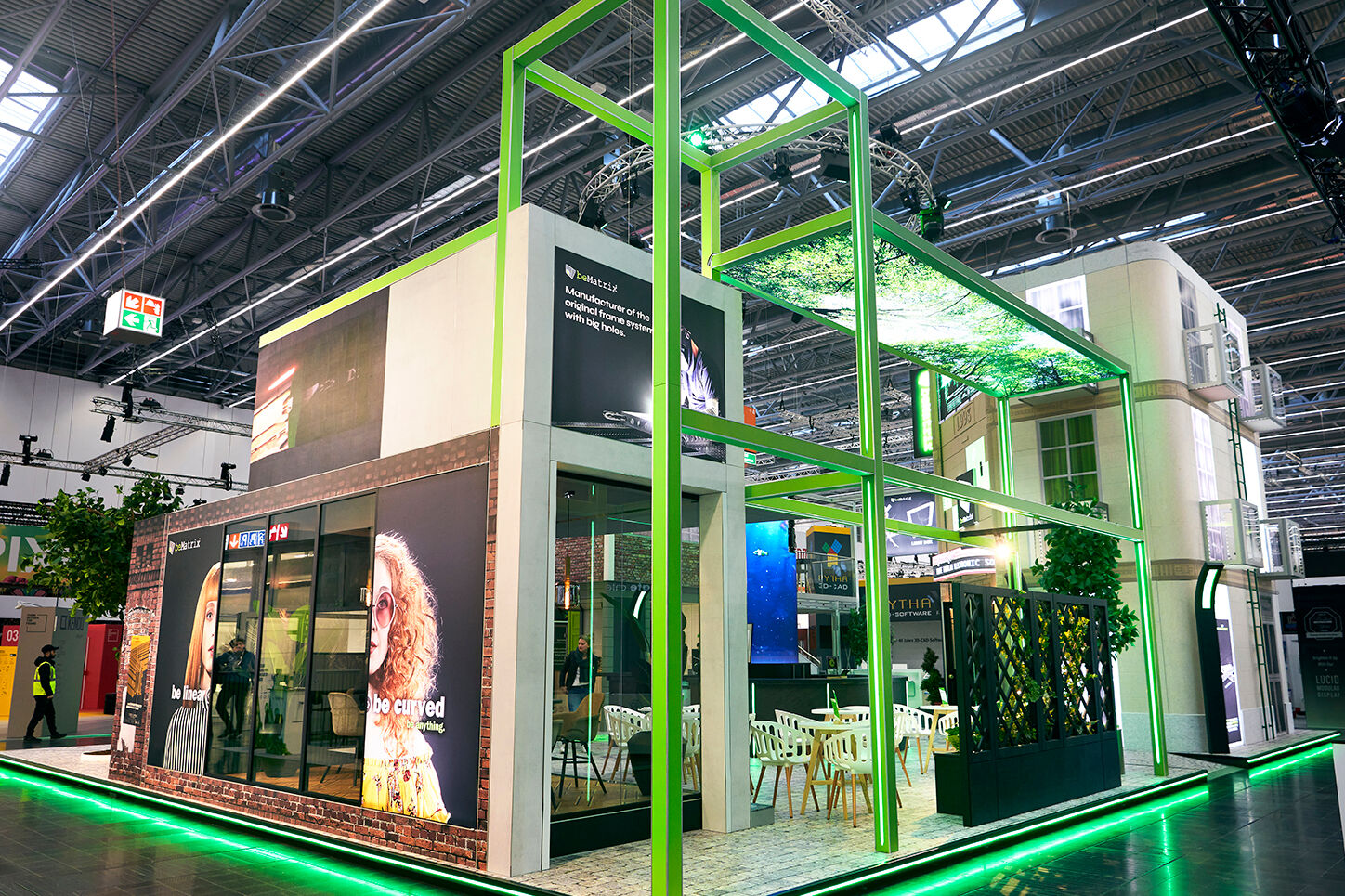 A modern exhibition booth with green neon lighting, glass walls, and digital screens, featuring seating areas and overhead greenery, set inside a large, industrial convention hall.