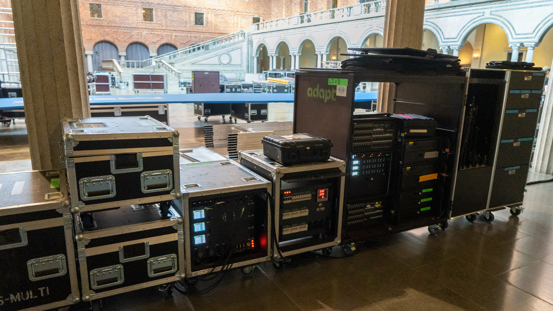 Stacked audio and technical equipment cases on wheels are set up in a large hall with high ceilings, arched windows, and visible staircases in the background, suggesting event setup or preparation.