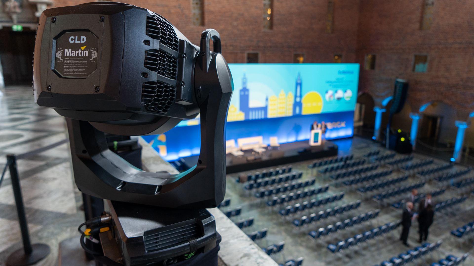 A close-up of a stage spotlight overlooks a large, empty auditorium with rows of chairs facing a colorful conference stage displaying cityscape graphics and a podium. Two people stand near the stage.