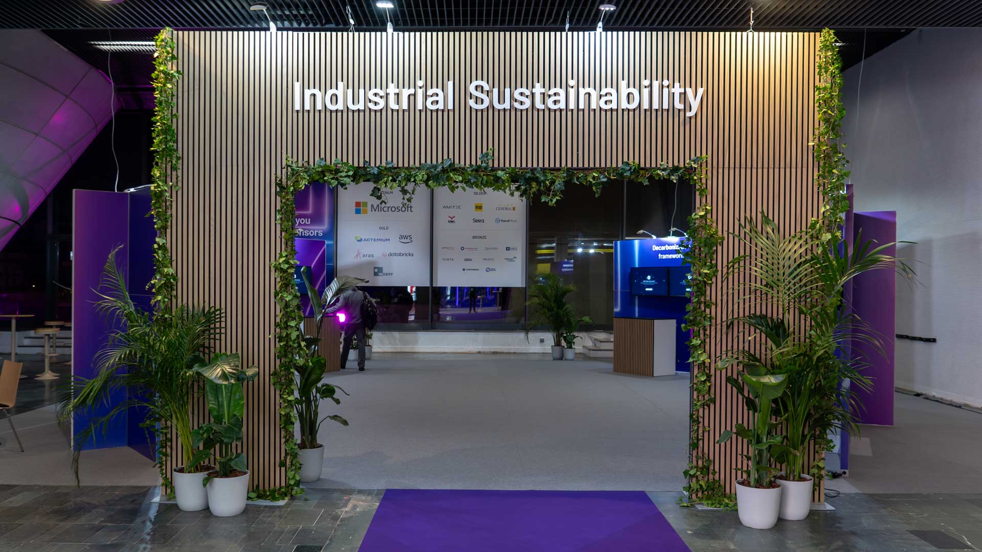 Entrance to an event space with a sign reading Industrial Sustainability, decorated with green plants and wooden slats. Several sponsor logos are visible inside on a white panel. The area is lit with purple and white lights.