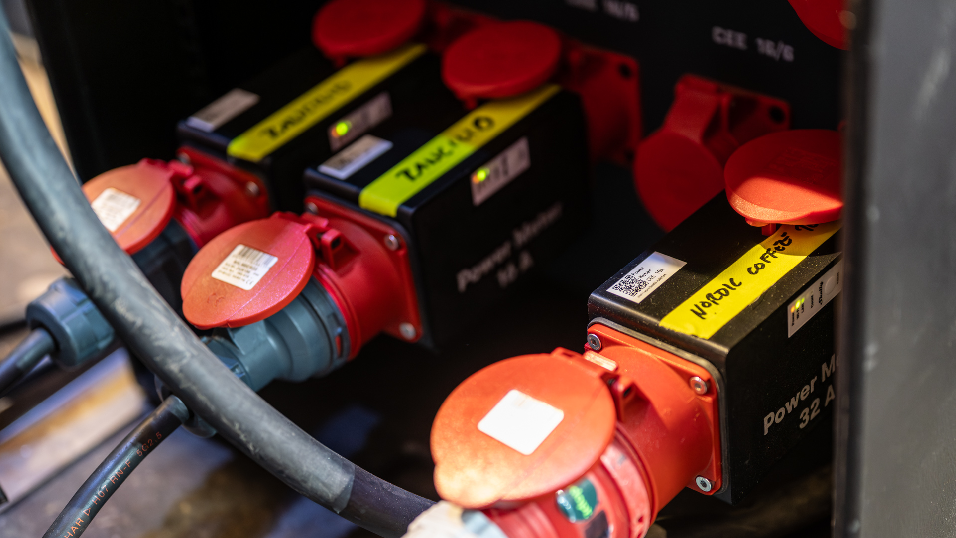 Close-up of industrial power outlets with red covers and labeled boxes, connected by thick black cables inside an electrical panel. The labels read Power Meter 32A and some text is handwritten on yellow tape.