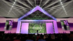 A speaker stands on a large, futuristic stage with purple lighting, geometric shapes, and screens displaying Shaping a sustainable future across industries to an audience. Two vertical screens show the speakers image.