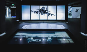 A dark exhibition space features a large screen showing a fighter jet in flight. In the foreground, another screen displays two people operating equipment in a control room.