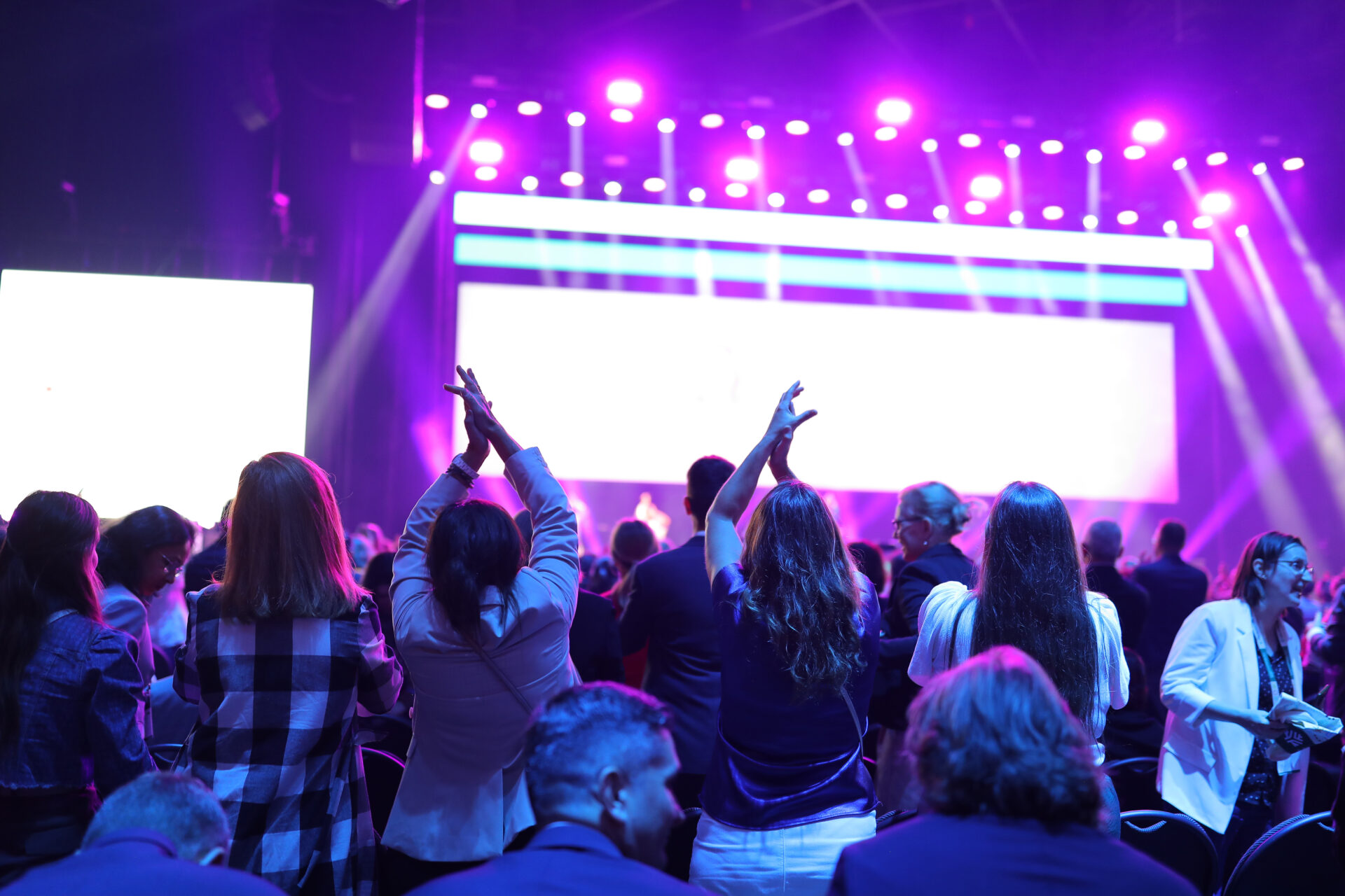 A crowd of people stands and cheers at an indoor event with bright stage lights and large screens in the background, creating an energetic and lively atmosphere.