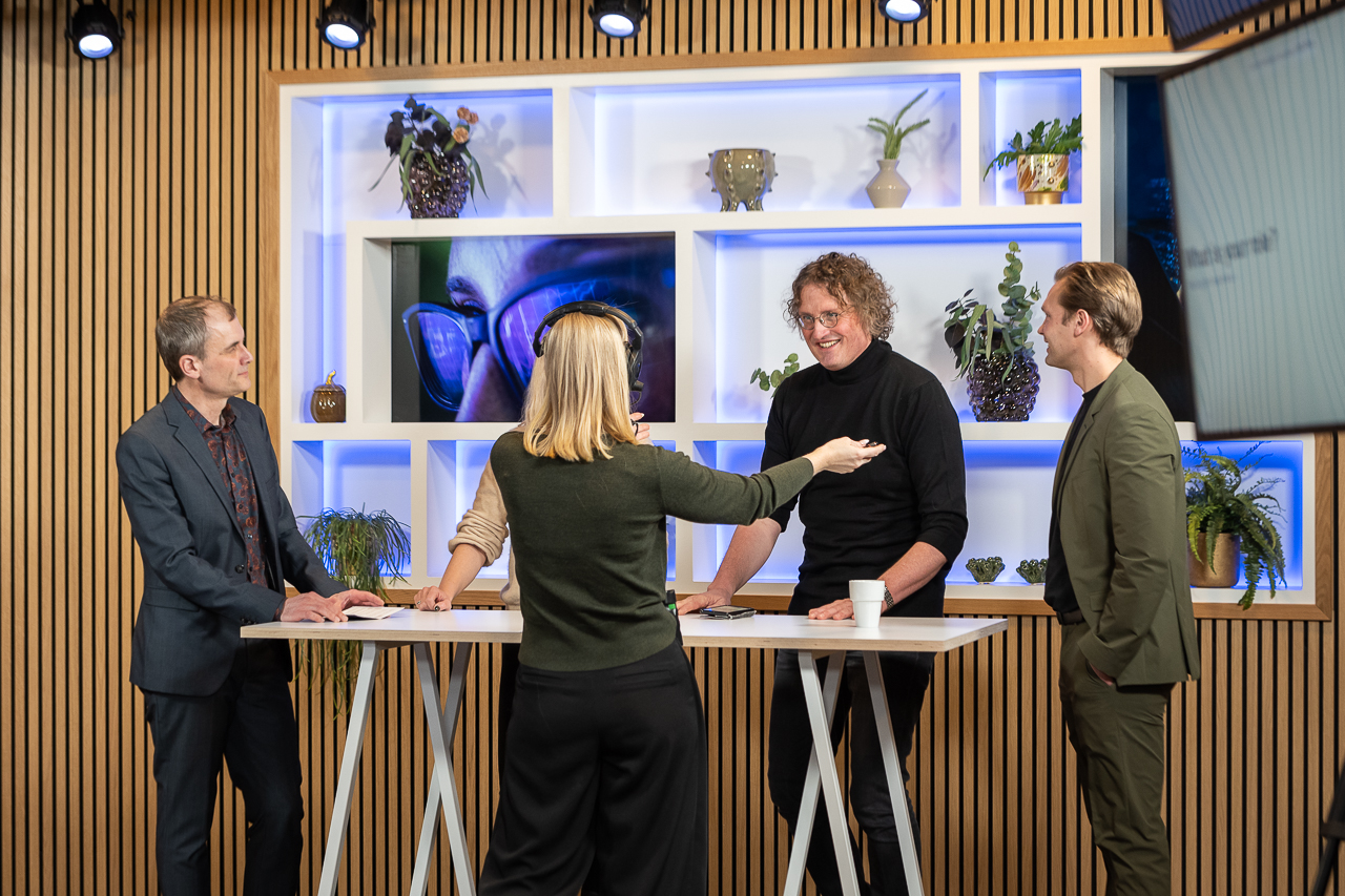 Four people stand around two white tables in a modern studio with wooden panel walls and plants on shelves. One woman gestures while talking to three men, and everyone appears engaged in conversation.