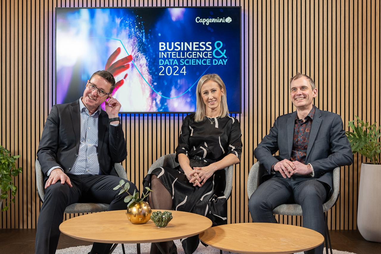 Three people in business attire sit smiling on chairs in front of a screen displaying Capgemini Business & Intelligence Data Science Day 2024, with wooden panel walls and plants as decor.