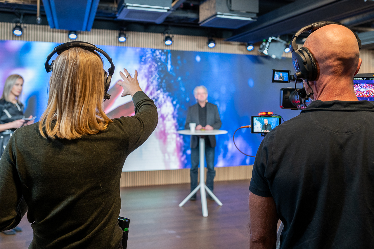 A woman with a headset gestures toward a man standing at a round table on a stage, while a cameraman films them; colorful lights illuminate the background.